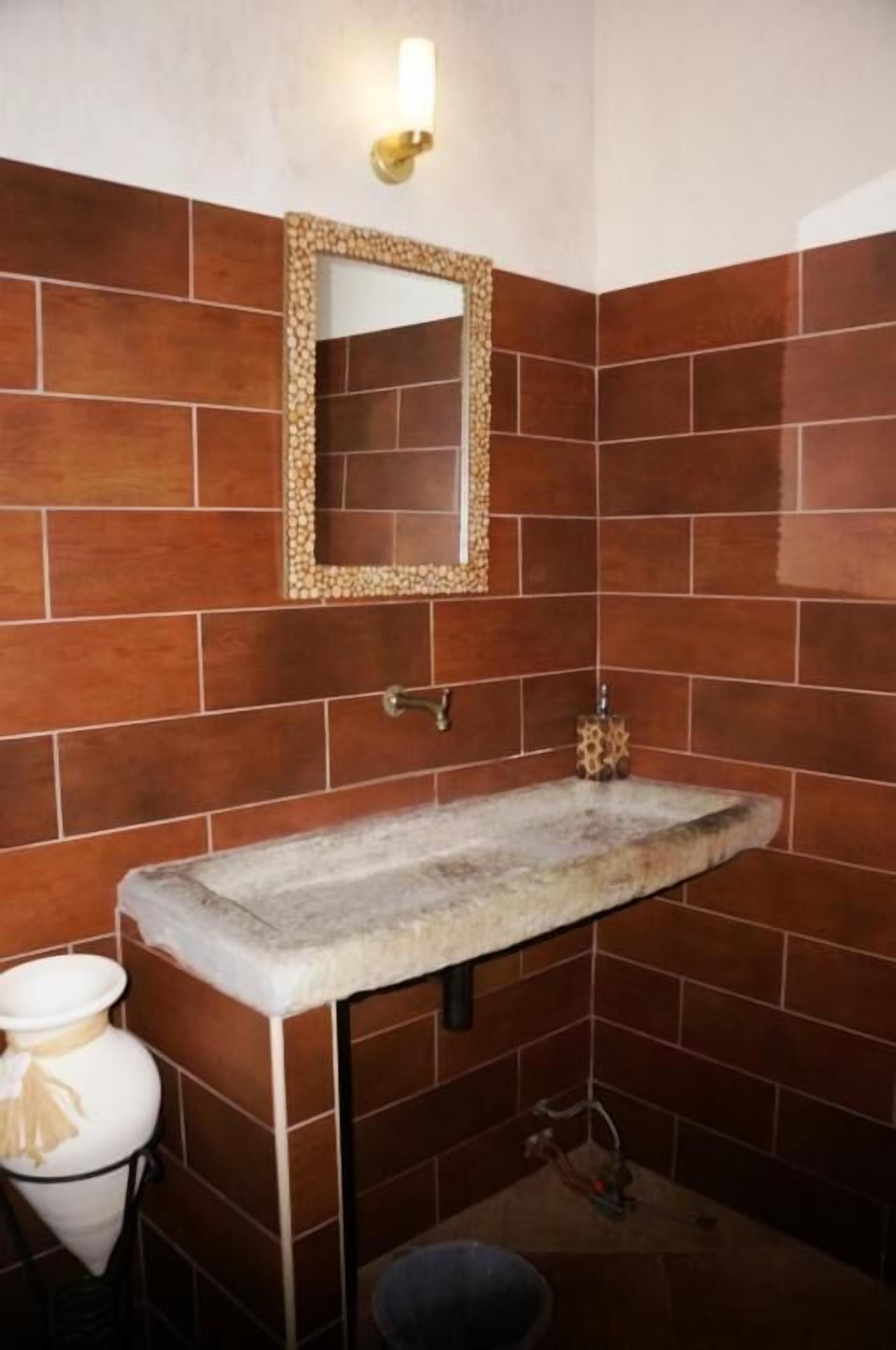 A bathroom with a stone sink, a mirror with a decorative frame, and brown tiled walls.