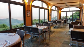 Un restaurante con mesas y sillas dispuestas para comer, ventanas amplias que ofrecen vistas del paisaje, y suelo de piedra.