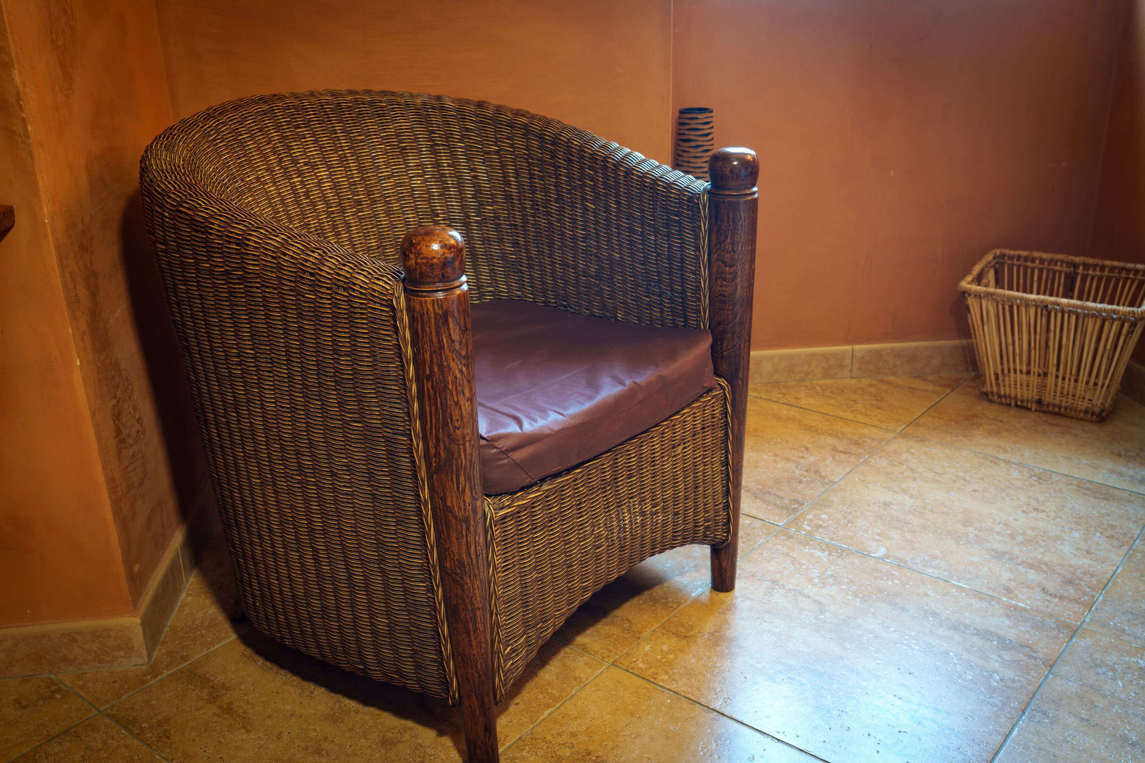 Una sedia in vimini con seduta imbottita e braccioli in legno, in una stanza con pavimento piastrellato.