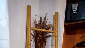 Una esquina con un toallero de bambú que sostiene dos toallas blancas, un florero con plantas secas y un estante de madera con un objeto negro encima.