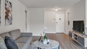 Un salon moderne avec un canapé gris, une table basse ronde agrémentée d’objets décoratifs, un téléviseur à écran plat posé sur un meuble en bois, et une porte blanche en arrière-plan.