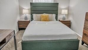 Ein ordentlich eingerichtetes Schlafzimmer mit einem grünen, mit Stoff bezogenen Bett, weißer Bettwäsche und zwei Nachttischen mit Lampen.