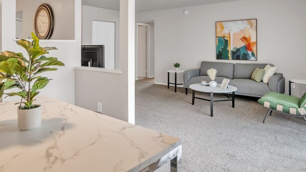 Ein modernes Wohnzimmer mit einem grauen Sofa, einem Couchtisch und einem grünen Sessel. Auf einem Marmortisch steht eine Pflanze und an der Wand hängt ein Bild.
