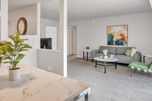 Una sala de estar moderna con un sofá gris, una mesita de centro y un sillón verde. Hay una planta sobre una mesa de mármol y un cuadro en la pared.