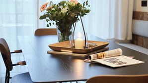 Une table de salle à manger en bois, ornée d’un vase de fleurs, d’une bougie et d’un plateau sur lequel repose un magazine enroulé.