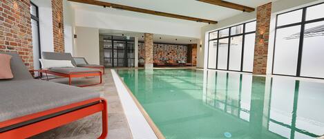 An indoor swimming pool with a modern design, featuring a red-framed lounge area and large windows.