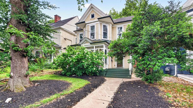 A two-story house with a front porch, a driveway, and a large tree in the foreground.
