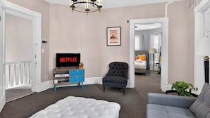 A room with a tufted white sofa, a grey armchair, a TV displaying 'NETFLIX,' a framed poster, and a chandelier.