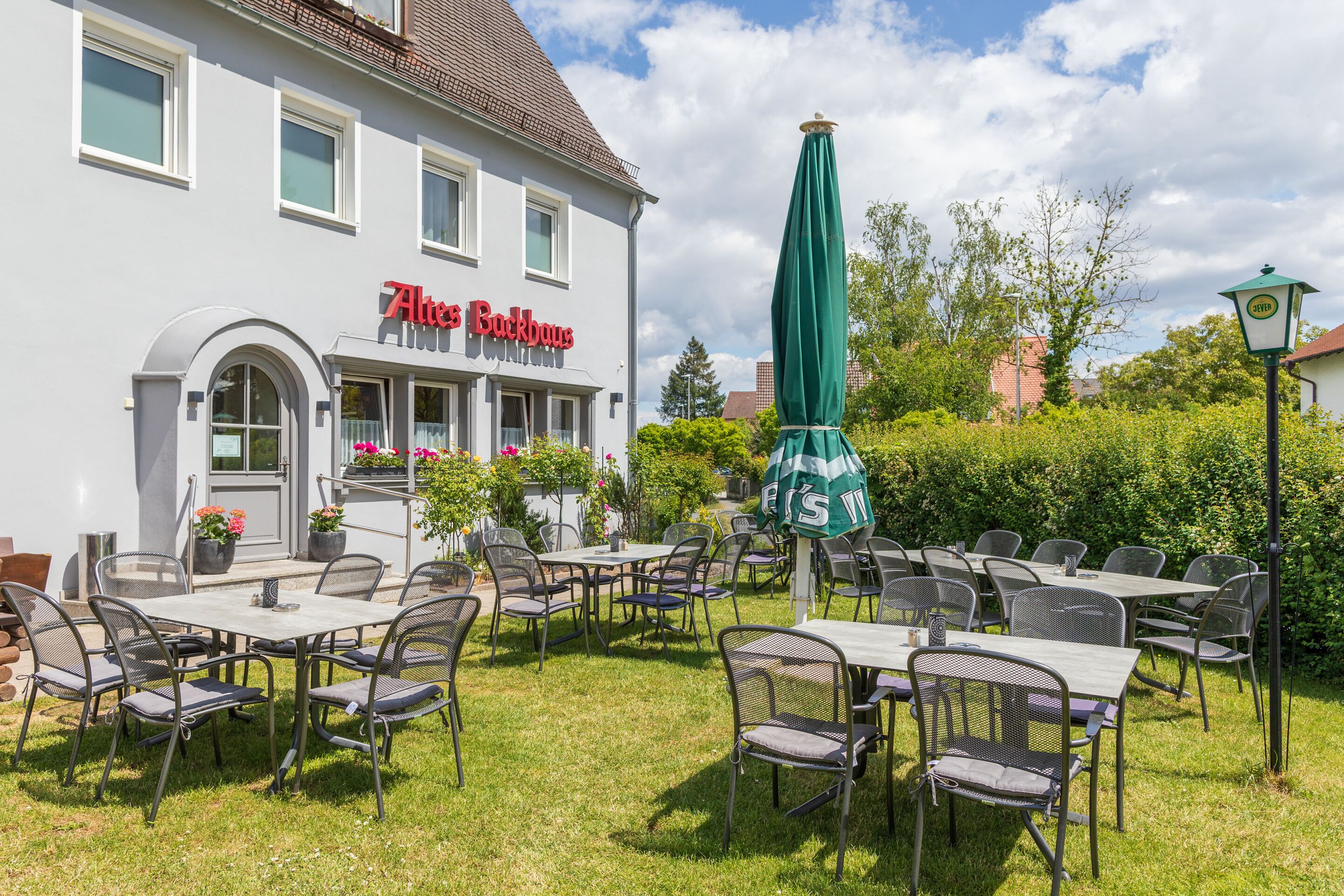 Un espace extérieur aménagé avec des tables et des chaises, un parasol vert et un bâtiment sur lequel on peut lire l’inscription « Altes Backhaus ».