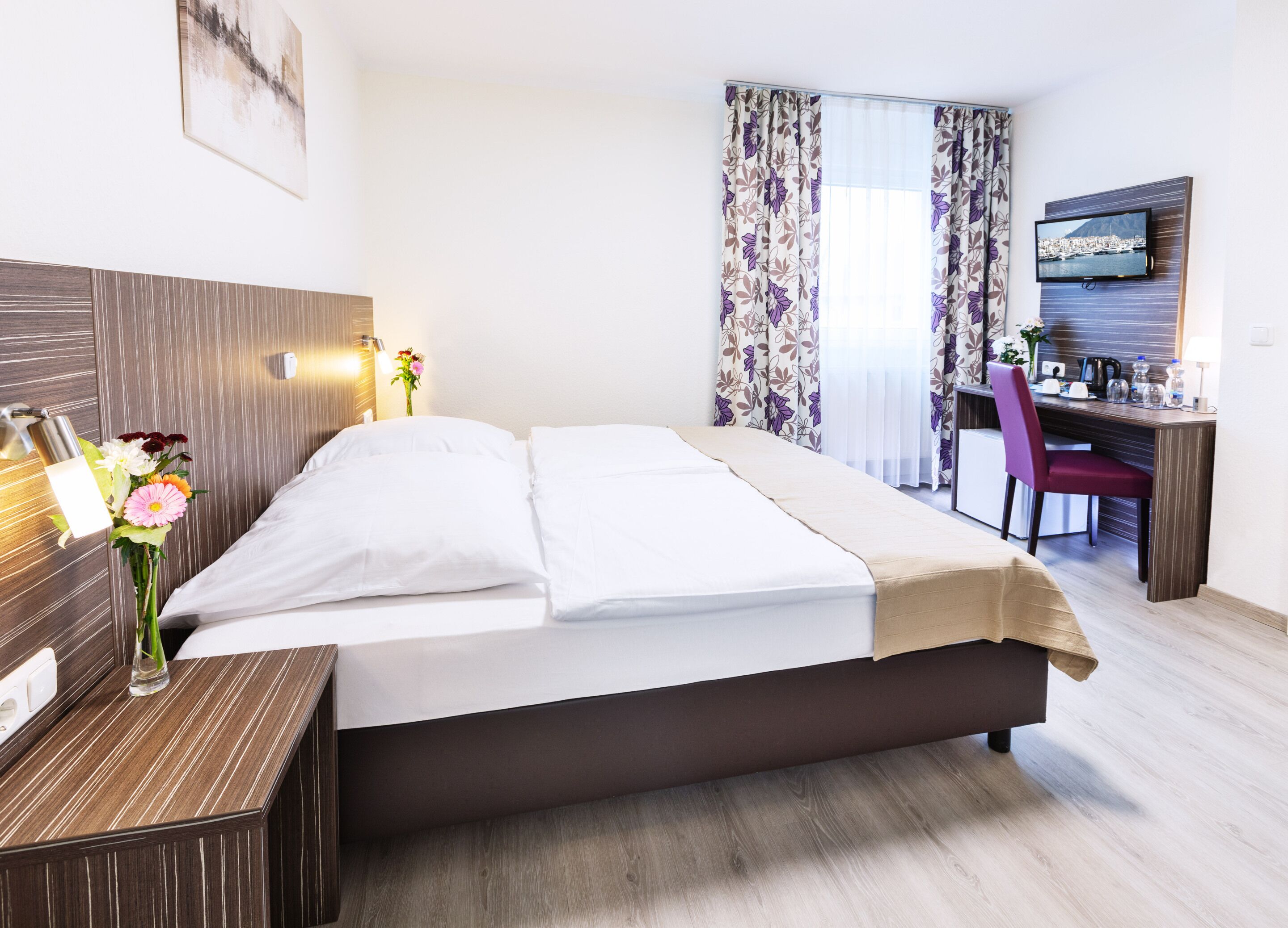 Een hotelkamer met een bed, nachtkastje, lamp, bloemen, een bureau met stoel, een televisie en een raam met gordijnen.