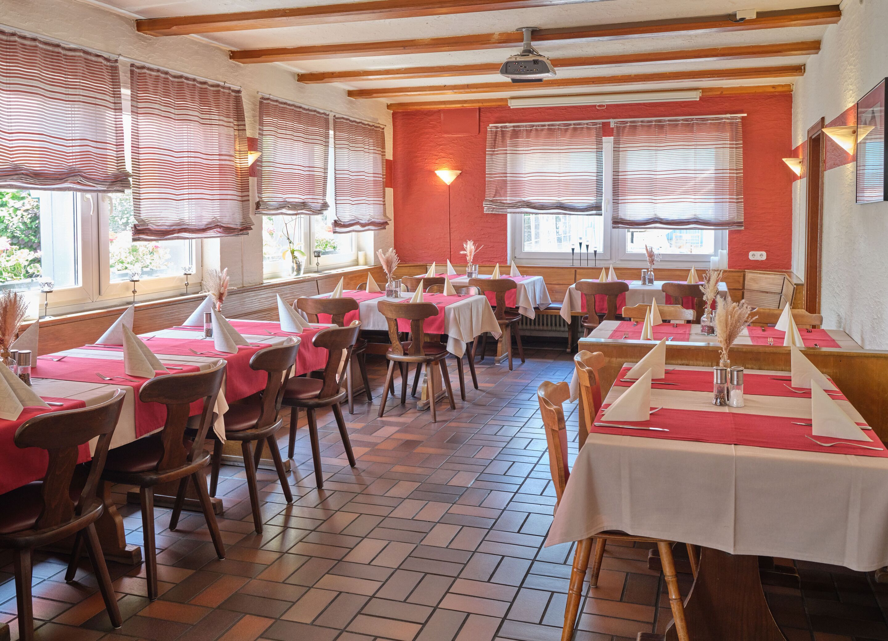 A restaurant with multiple tables and chairs, wooden beams on the ceiling, and large windows with striped curtains.