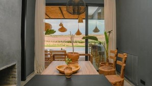Ein Essbereich mit Tisch für zwei Personen, Holzstühlen, einer Hängelampe und Blick auf eine natürliche Landschaft durch große Fenster.