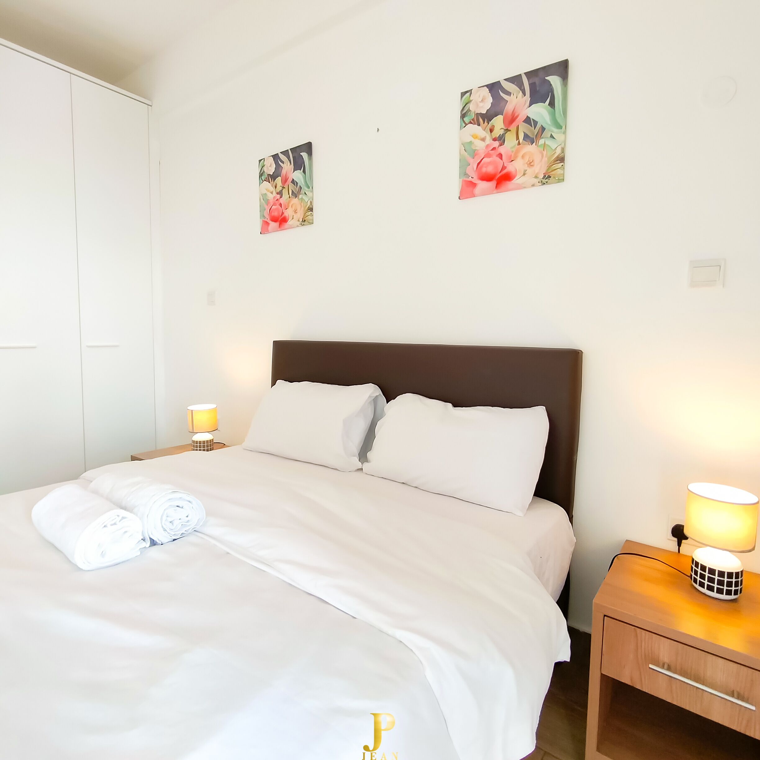 A neatly made bed with white linens, two wall-mounted paintings, and a bedside lamp.