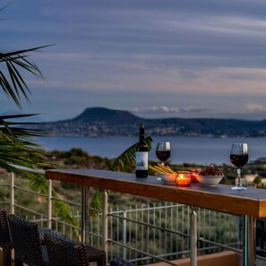 A balcony with a wooden table, a bottle of wine, two glasses, a bowl of grapes, and a candle. - Villa Roula 3BR Seaview Villa in Chania (Chania)