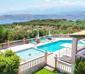 A pool area with a view of the sea and mountains, surrounded by greenery and a fence. - Villa Roula With Private Pool (Chania)