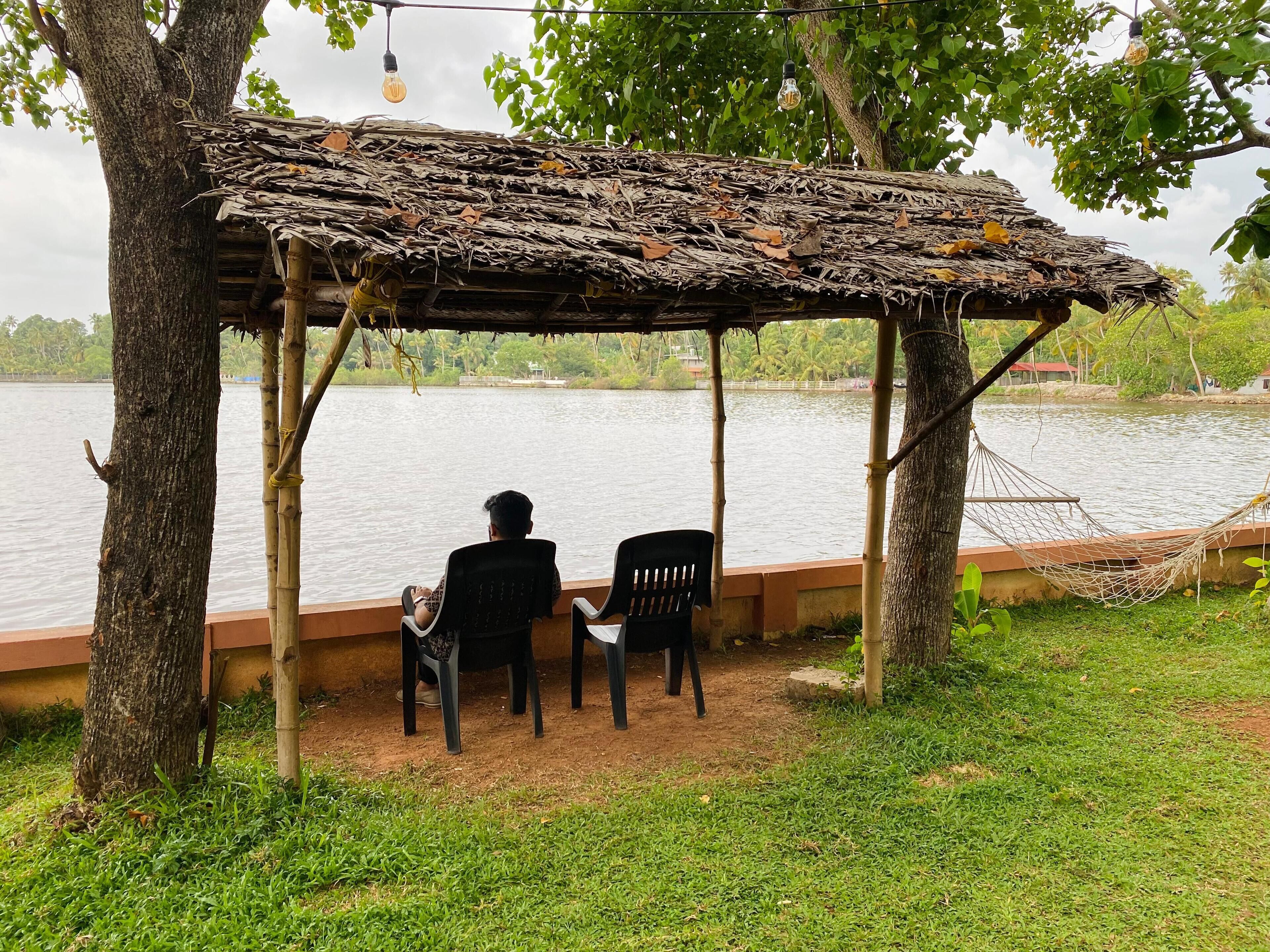 Un espace extérieur rustique, aménagé avec un toit de chaume, deux chaises et un hamac, situé au bord d’un lac.
