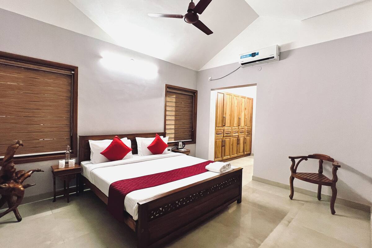 A bedroom with a bed, a ceiling fan, an air conditioning unit, wooden furniture, and a window with blinds.