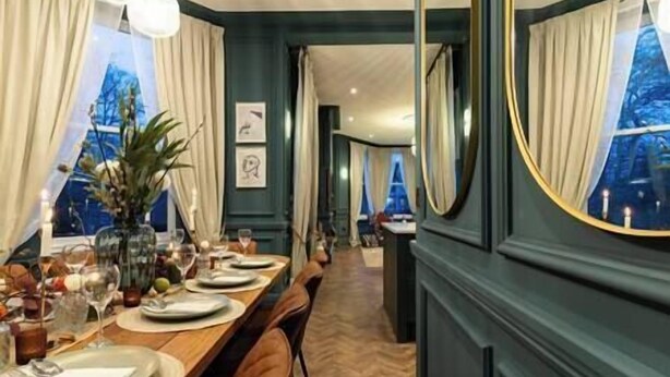 A dining area with a long table set for a meal, flanked by large windows with curtains. - Hob Hurst - Spacious 4 Bed Apt Hot Tub & Parking (Buxton)