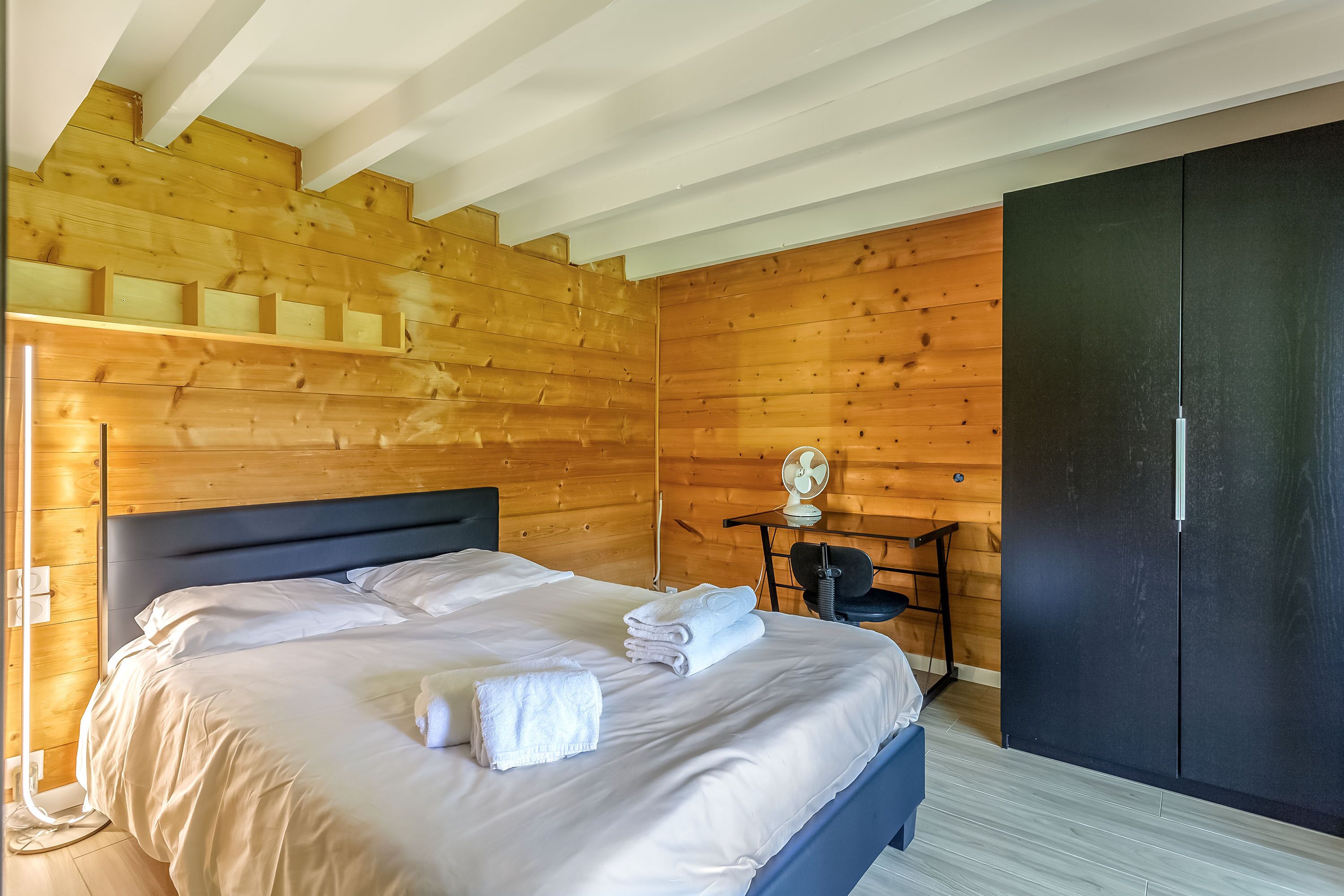 Un dormitorio con techo de madera, una cama con ropa de cama blanca, un armario negro y una mesita con un ventilador.