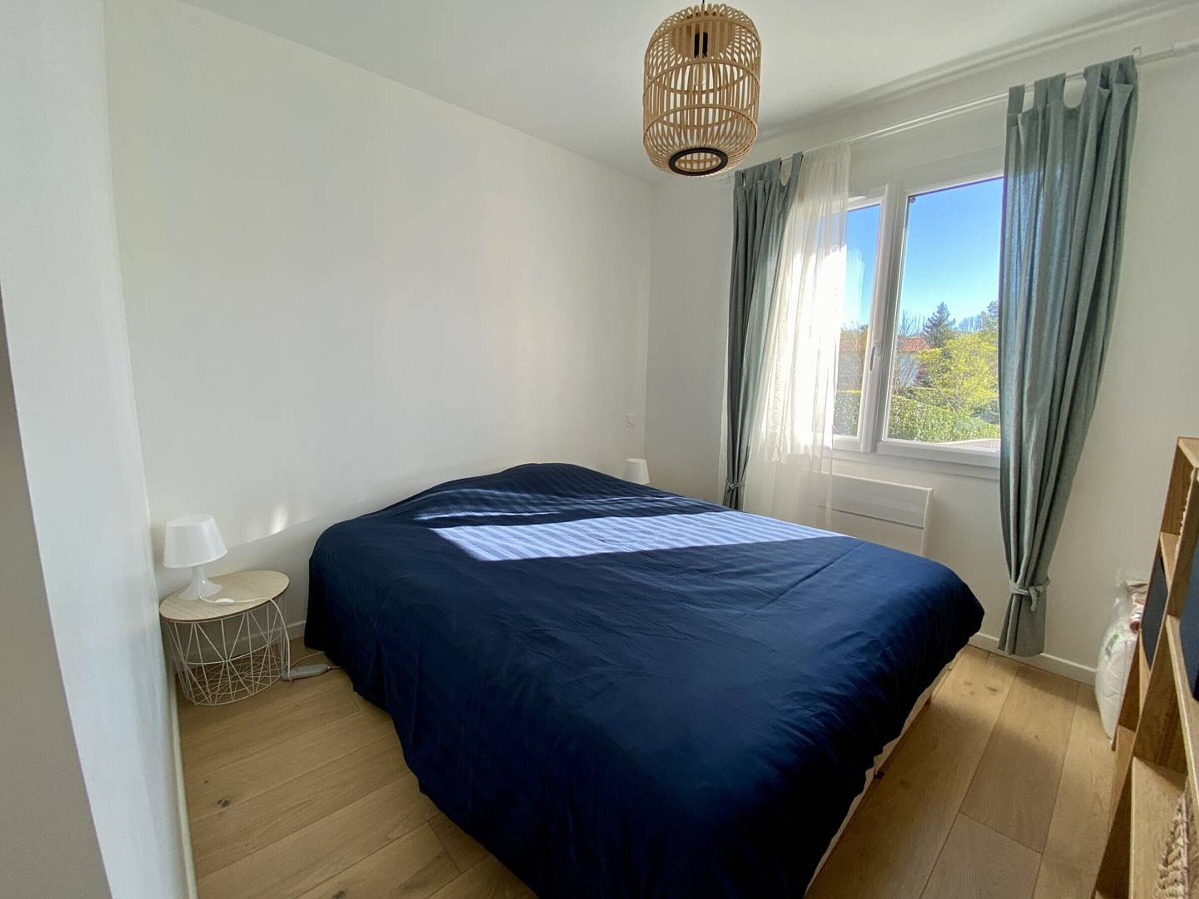 Een slaapkamer met een groot bed, een nachtkastje met een lamp, een raam met gordijnen en een hanglamp.