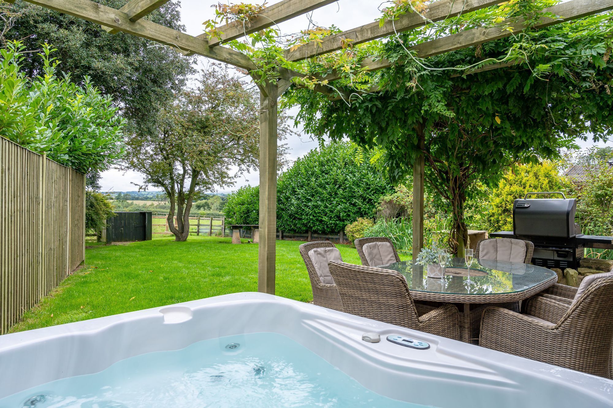 Patio trasero con jacuzzi, sillas de mimbre, mesa de vidrio, parrilla y pérgola con plantas trepadoras.