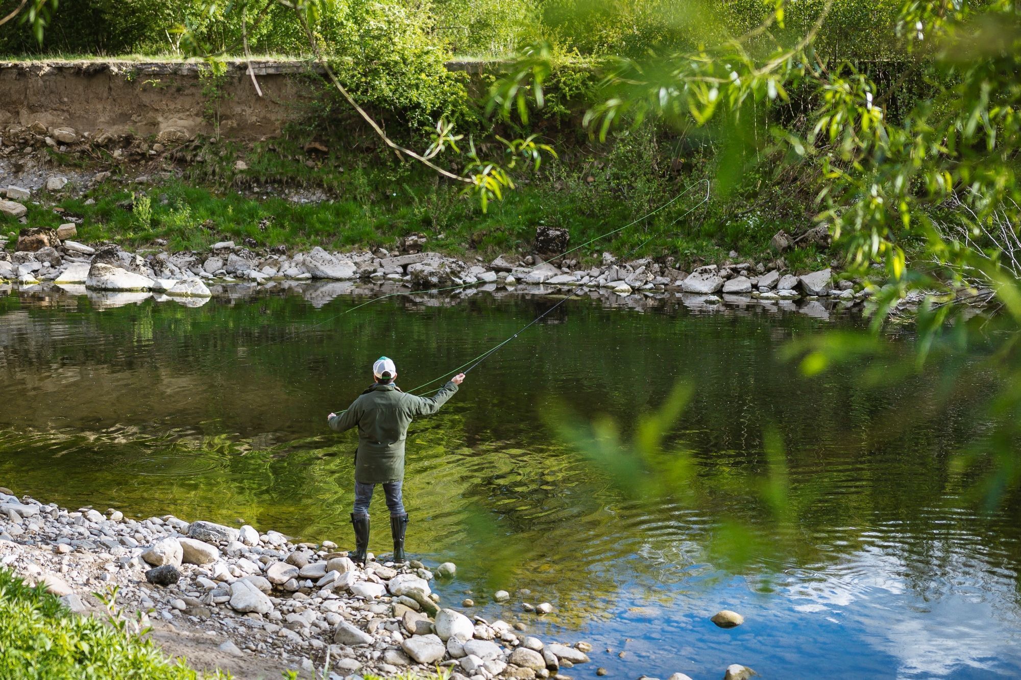 A person fishing by a river with a rocky shore and dense greenery in the background.
