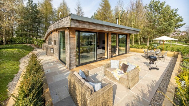 Een modern huis met een gebogen dak, grote glasramen en een terras met rieten meubilair.