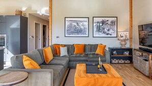 Un salon moderne avec un canapé d’angle gris, des coussins orange, une table basse en bois et un téléviseur à écran plat. Deux photographies encadrées sont accrochées au mur et un panier en osier est posé sur le sol.
