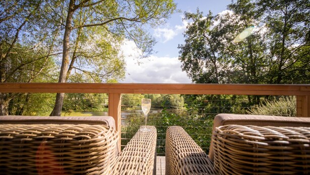 Des chaises en osier sont disposées face à une terrasse en bois, offrant une vue sur un lac et des arbres.