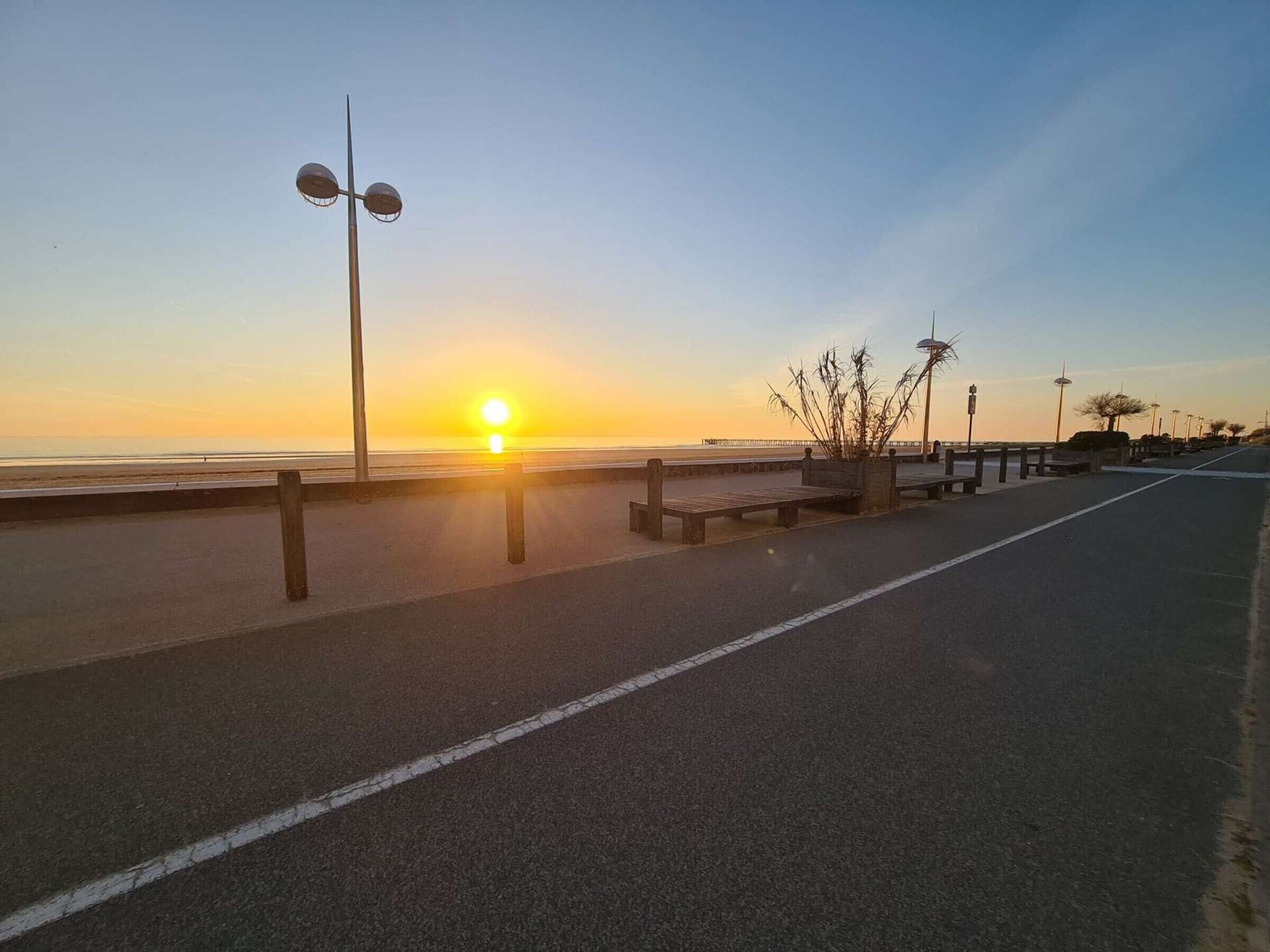 A coastal road with a sunset view, benches, street lamps, and a clear sky.