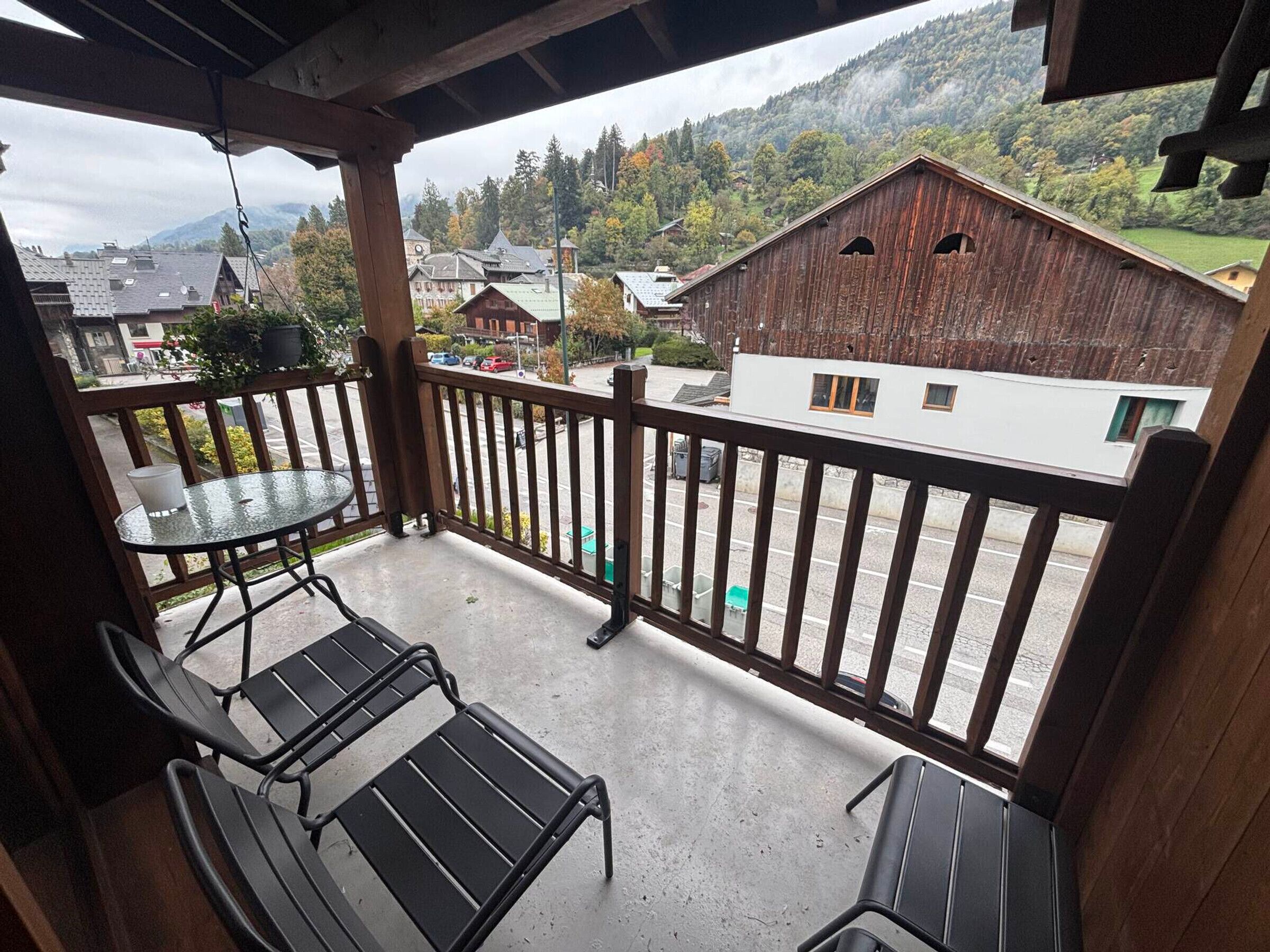 Een balkon met twee metalen stoelen en een glazen tafel, met uitzicht op een straat met gebouwen en een berg op de achtergrond.
