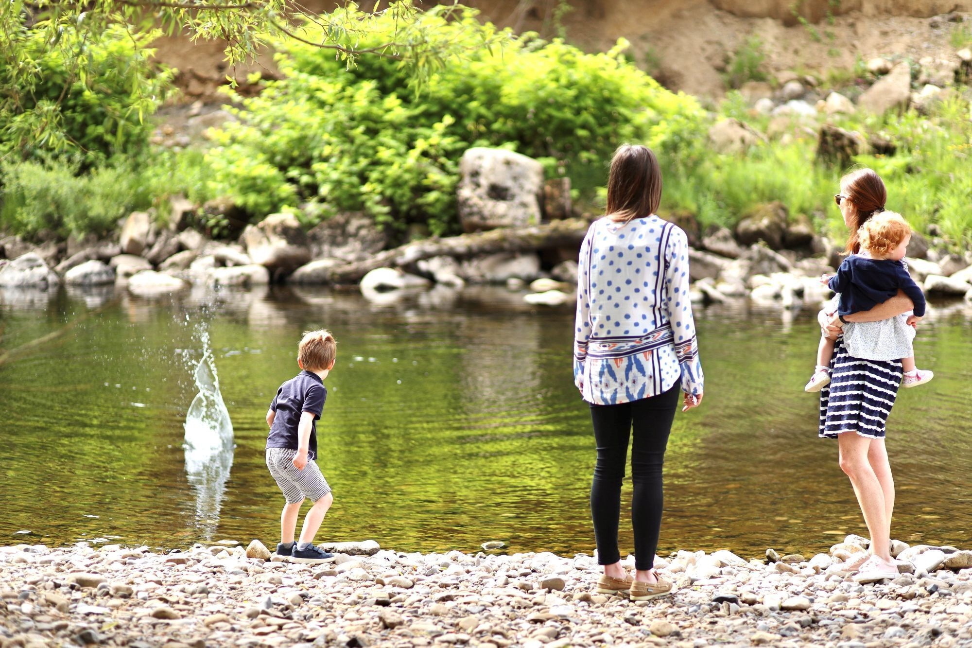 Una familia junto a un río tranquilo, con rocas y vegetación al fondo.