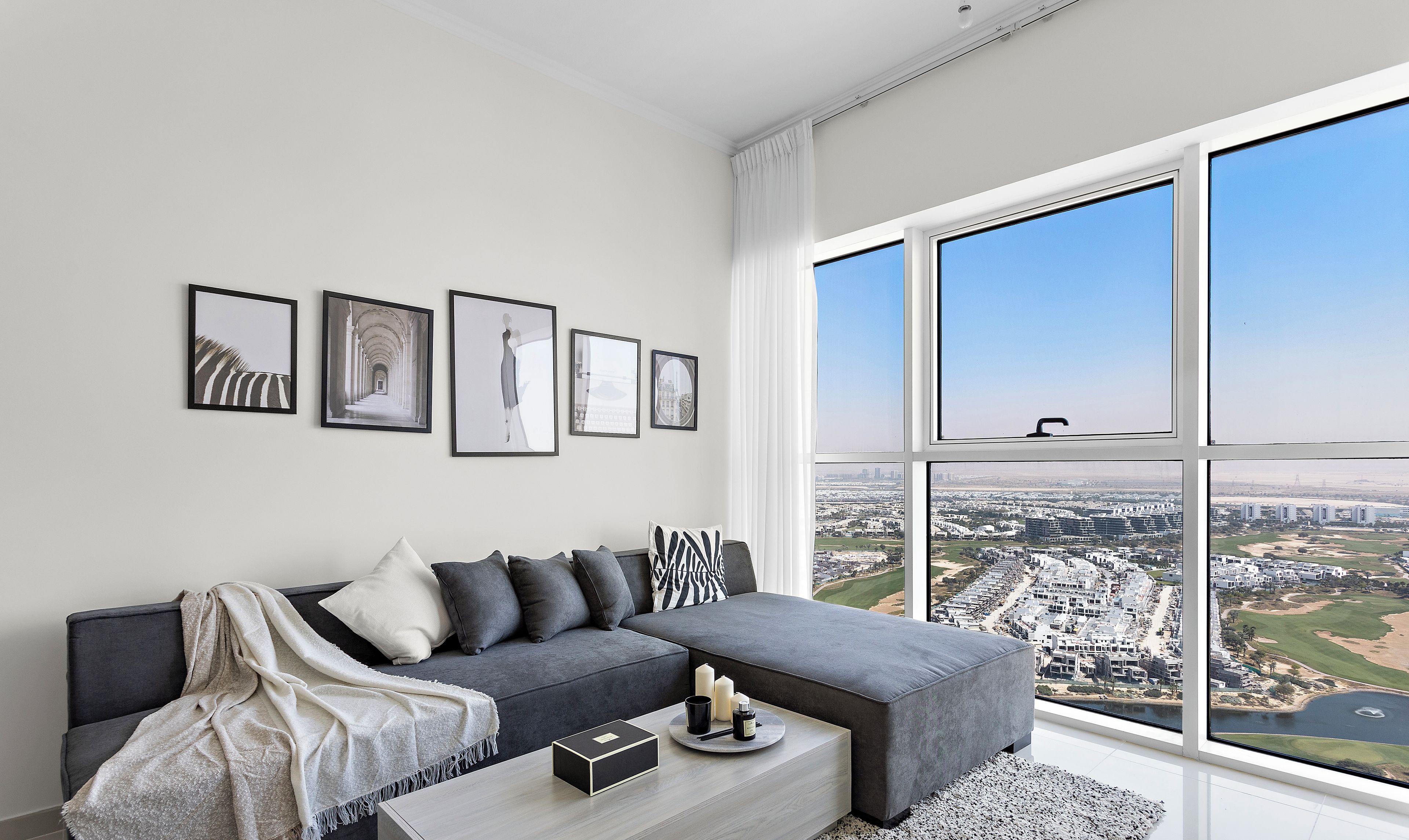A modern living room with a grey sectional sofa, a coffee table with decorative items, and framed artwork on the wall. A large window offers a view of a cityscape.