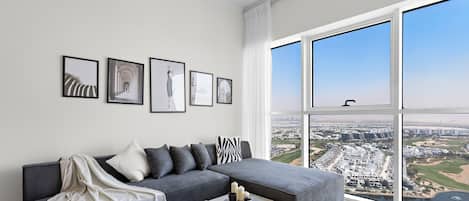 Un salon moderne avec un canapé d’angle gris, une table basse agrémentée d’objets décoratifs et des œuvres d’art encadrées accrochées au mur. Une grande fenêtre offre une vue sur le paysage urbain.