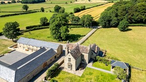 A large stone building with a flat roof, surrounded by green fields and trees. - Hollins Farmhouse -  a house that sleeps 10 guests  in 5 bedrooms (Leyburn)