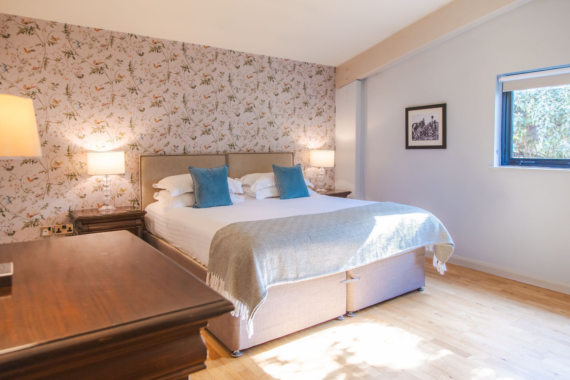 A bedroom with a large bed, two bedside lamps, a wooden nightstand, a dresser, and a window with a view of greenery.