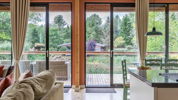A modern interior with a sofa, dining table, and chairs. There is a large window with curtains, offering a view of a deck and trees. - Cara -  a lodge that sleeps 6 guests  in 3 bedrooms (Richmond)