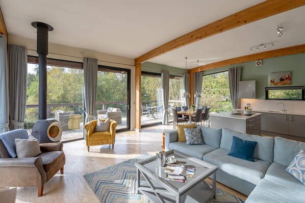 Una sala de estar moderna con un sofá, un sillón y una mesa de centro. Hay un balcón con vistas a árboles y una cocina con un fregadero y armarios.