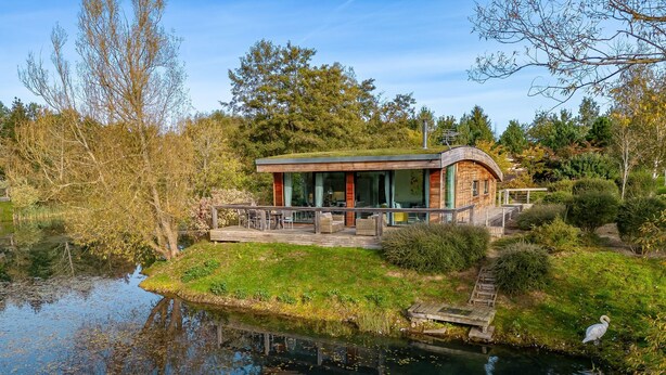 Una casa moderna con un balcón que da a un estanque, rodeada de árboles y vegetación.