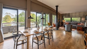 Une pièce spacieuse comprenant un coin repas, un canapé et une grande fenêtre donnant sur des arbres et une étendue d’eau.