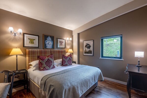 A bedroom with a large bed, two bedside lamps, a nightstand, a chair, and a desk with a lamp. There are framed pictures on the wall and a window with a view of greenery.