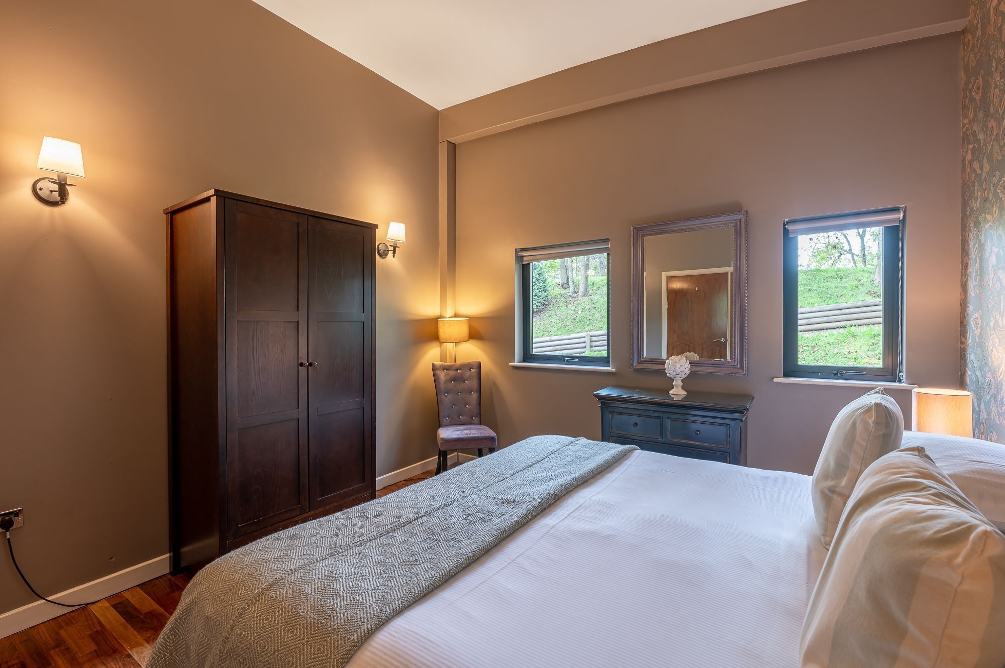 Une chambre dotée d’un grand lit, d’une armoire, d’une commode, de deux lampes fixées au mur et d’une fenêtre donnant sur un paysage verdoyant.