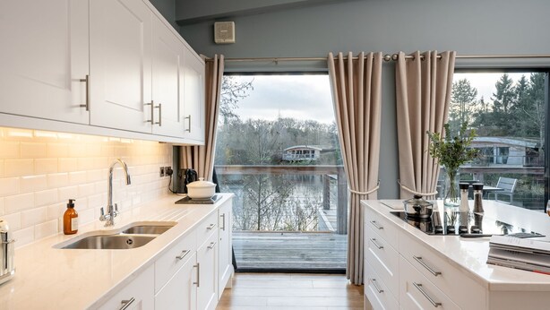 A modern kitchen with white cabinets, a large window with curtains, and a view of a house across a body of water. - Lola -  a lodge that sleeps 6 guests  in 3 bedrooms (Richmond)