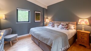A bedroom with a large bed, two bedside lamps, a chair, a wooden dresser, and a window with a view of greenery. - Juniper -  a lodge that sleeps 6 guests  in 3 bedrooms (Richmond)