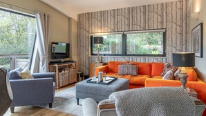 A living room with a grey sofa, an orange sofa, a television, and a large window with a view of trees. - Juniper -  a lodge that sleeps 6 guests  in 3 bedrooms (Richmond)