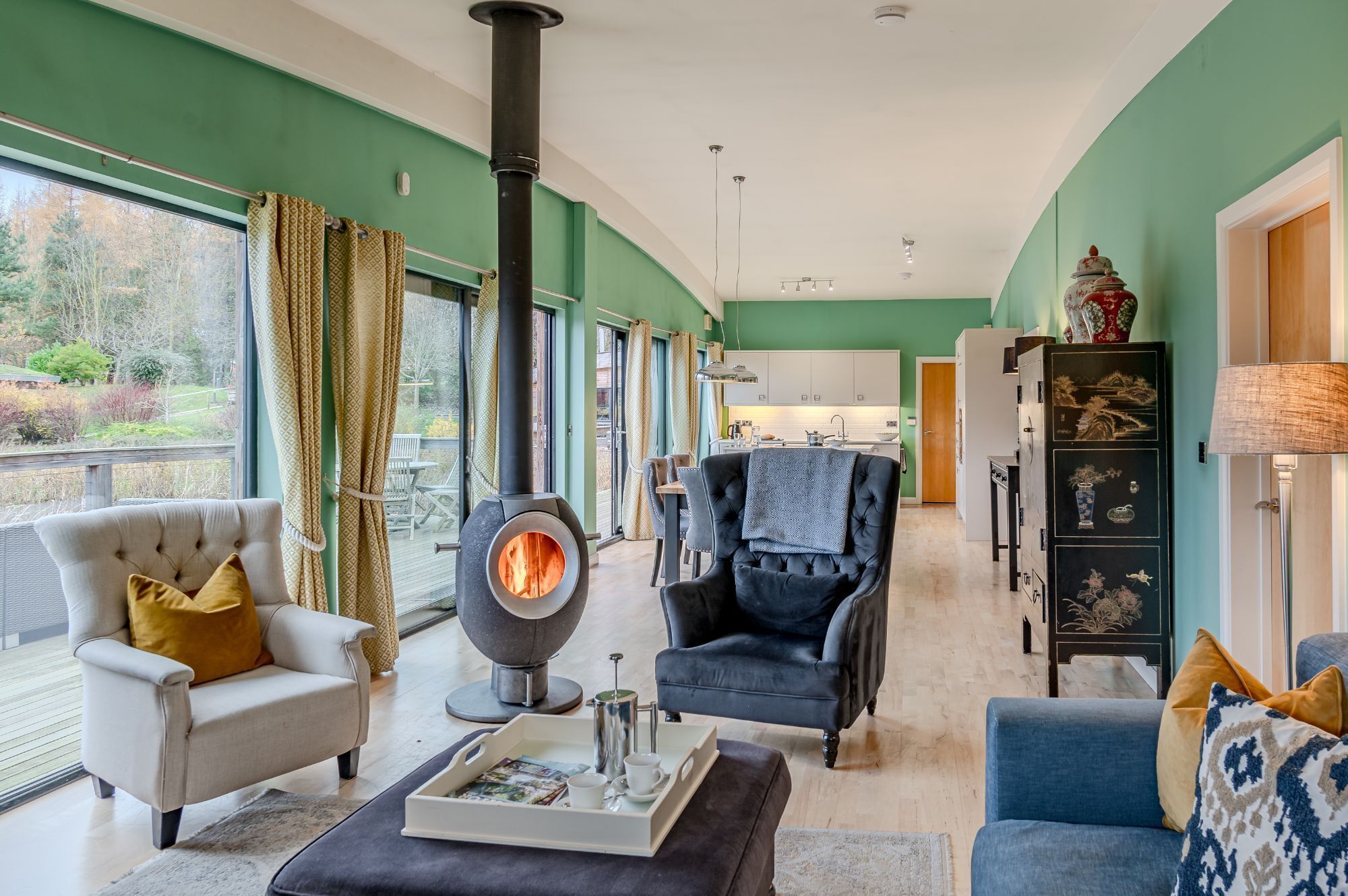 A living room with a wood-burning stove, a sofa, a chair, and a dining area with a kitchen in the background.