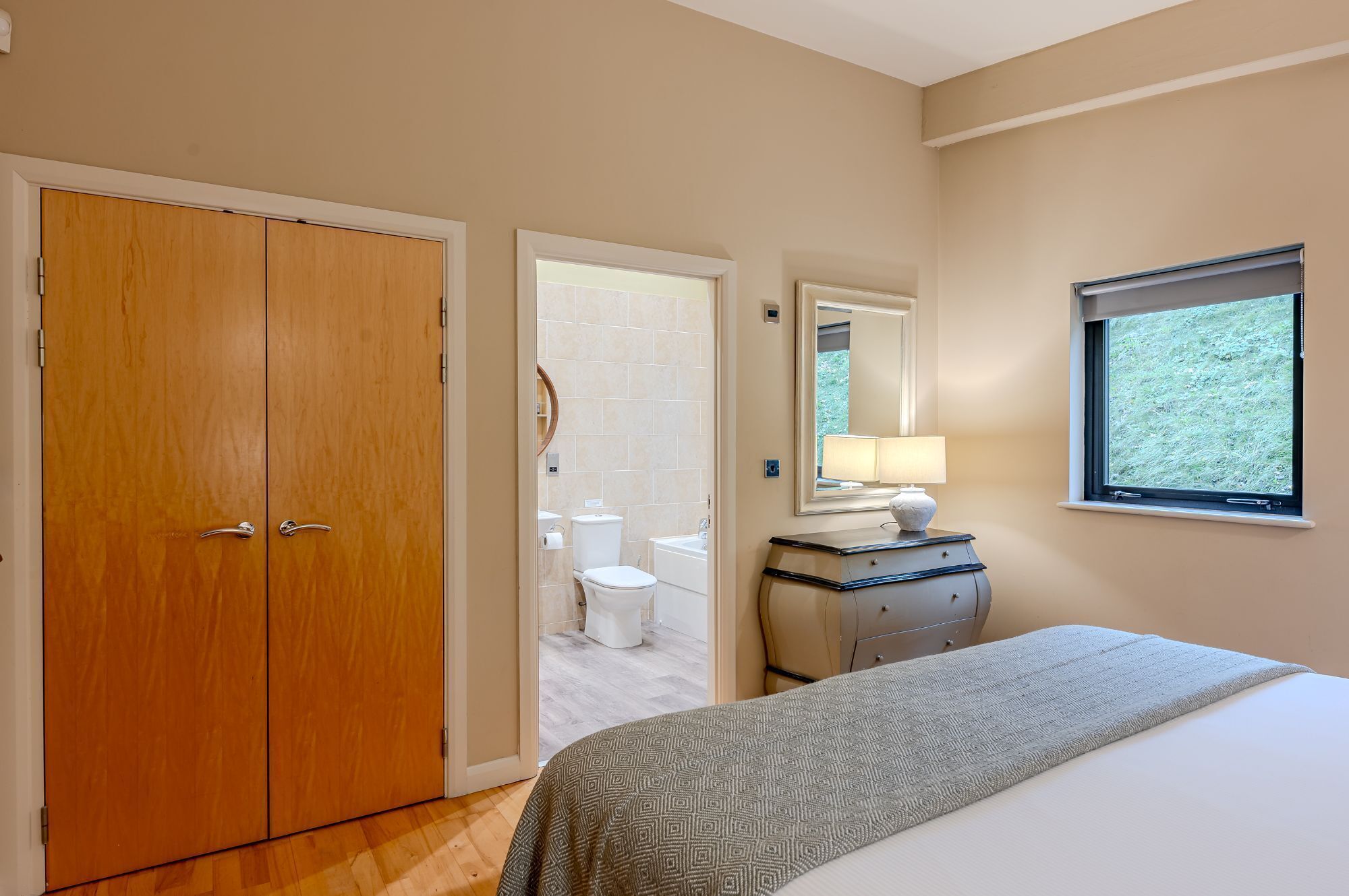 Quarto com cama, cômoda, janela e banheiro visível por uma porta aberta.