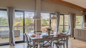A dining area with a wooden table, four chairs, and a vase of flowers. Large windows offer a view of a lake and trees.