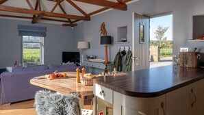 Un salon chaleureux avec un canapé, une table en bois sur laquelle se trouve de la nourriture, une lampe et un téléviseur. On y trouve également un coin cuisine avec un comptoir et des placards, et une vue sur un jardin par une porte ouverte.