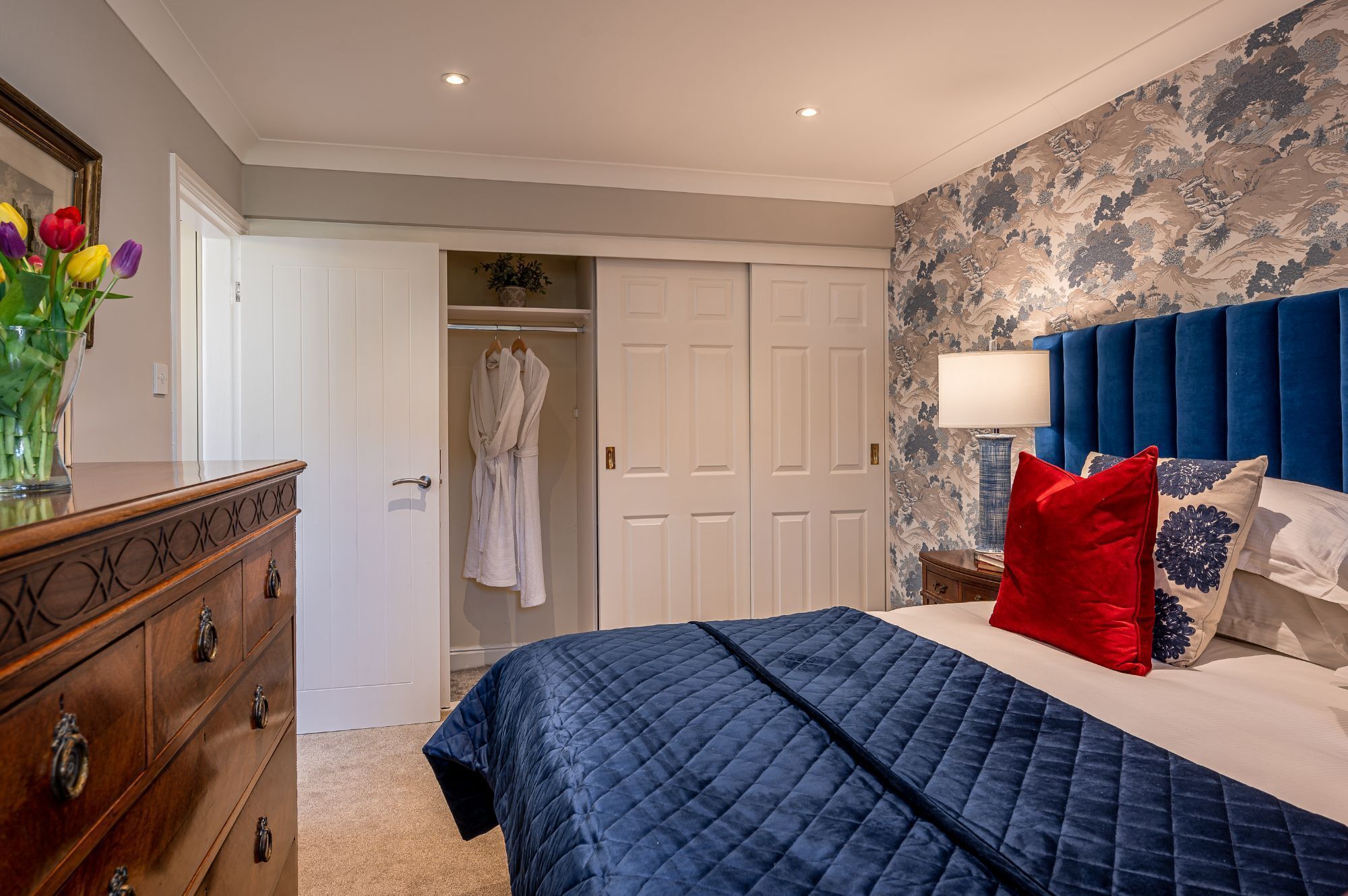 A bedroom with a bed, a dresser, a robe hanging on a door, and a nightstand with a lamp.